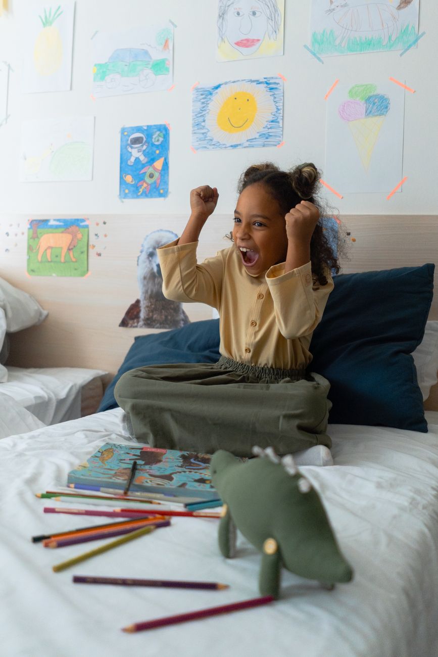 petite fille heureuse qui lève les bras, un livre est posé sur son lit